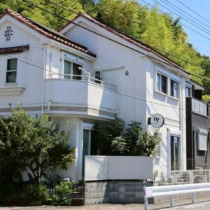 Two-story white guesthouse facade with balcony, small front greenery and bamboo backdrop, DP小山町Machida, Tokyo