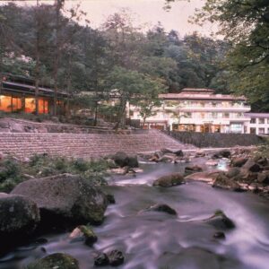 Dusk riverside scene at Gensenkan with lit ryokan buildings beside a rocky stream and forested banks in Tochigi