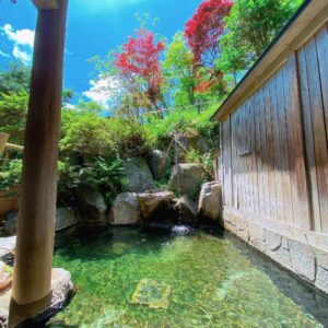 Clear outdoor bath with stone edge, wooden fence and red-leaf trees at Hakuunsou in Gifu