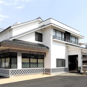 White two-story ryokan building with covered entrance, parking area and low hills in the background