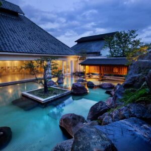 Outdoor onsen bath with rock garden and illuminated traditional buildings at dusk, Shiragiku, Ōita