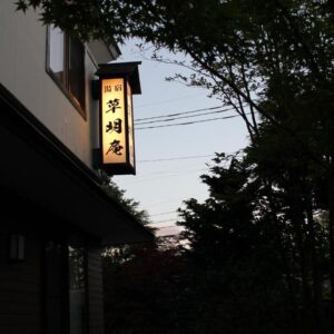 Dusk view of illuminated kanji sign and ryokan entrance framed by trees in Gunma