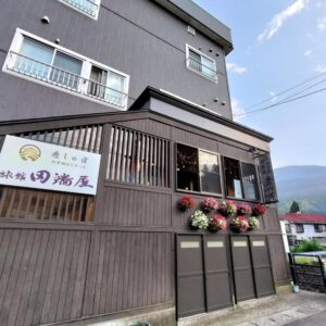 Wooden slatted ryokan facade with hanging flowers and signboard, mountain backdrop in Niigata's Chūbu region