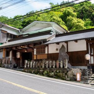 Traditional wooden ryokan exterior with lanterns and stone markers beside roadside and green hillside in Kanagawa