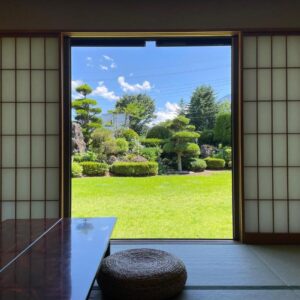 Tatami room with shoji screens, low table and round cushion opening onto a trimmed garden under blue sky in Yamanashi