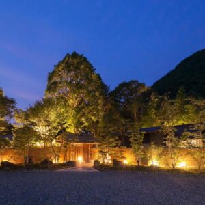 Dusk view of illuminated stone entrance and lanterns with trees and mountain backdrop at Ufufu Shizuoka