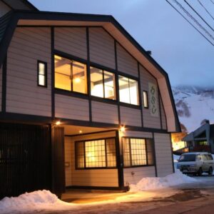 Two-story Warabiso exterior with illuminated windows, snow-covered street and parked cars in Niigata mountain town
