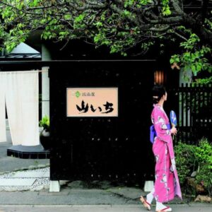 Person in pink yukata walking past Yamaichi entrance with noren and wooden gate in Iwate