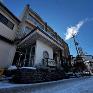 Snow-covered exterior of Ryokan Tsuruya with entrance and street under blue sky in Nagano, Chūbu