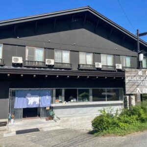 Two-story dark wooden ryokan facade with entrance, upper balconies and roadside greenery in Nagano, Chūbu
