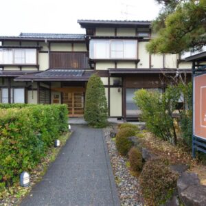 Walkway to traditional two-story ryokan entrance with wooden signboard, clipped shrubs and pine in Gifu, Chūbu region