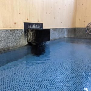 Indoor onsen bath with wooden spout, tiled floor and stone edging at Onsenya Hirosuke, Fukushima